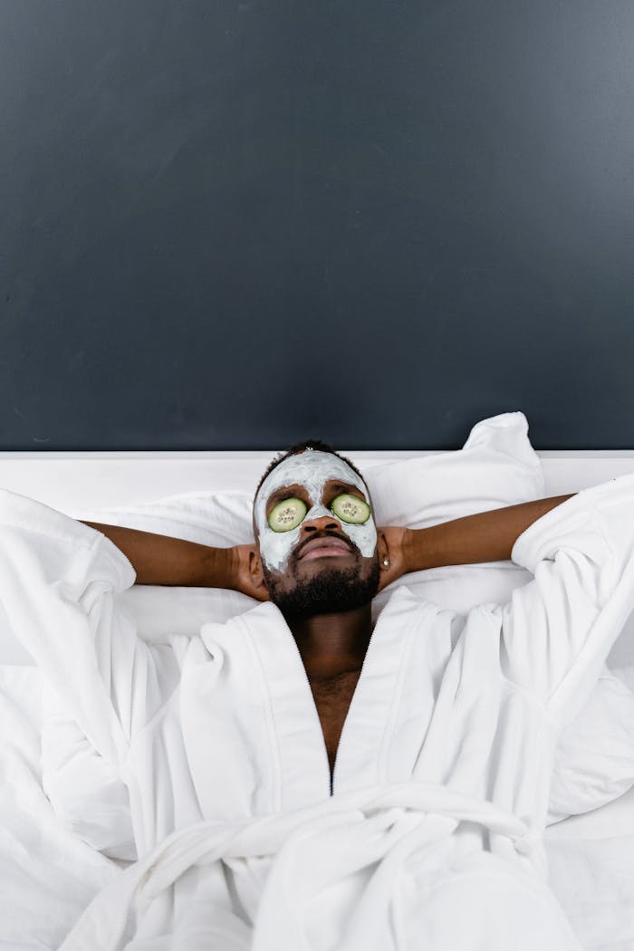 African American man in a white robe enjoying a cucumber facial mask, embodying relaxation and self care.